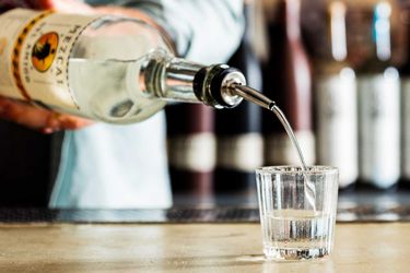 A glass of mezcal is poured from a bottle on a bar top.