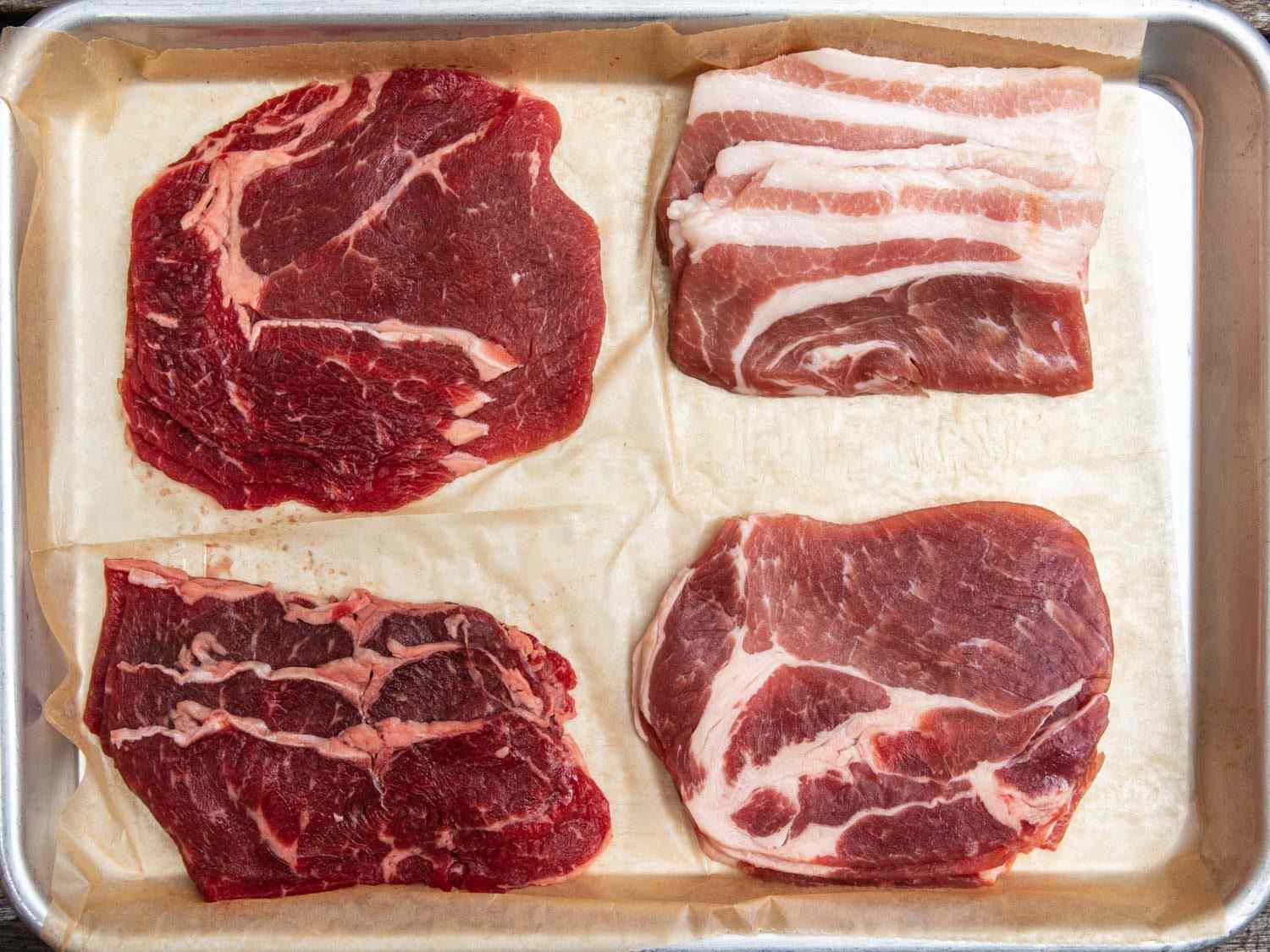 Different cuts of raw beef and pork for Korean barbecue, including thinly sliced ribeye and pork shoulder, and pieces of pork belly.