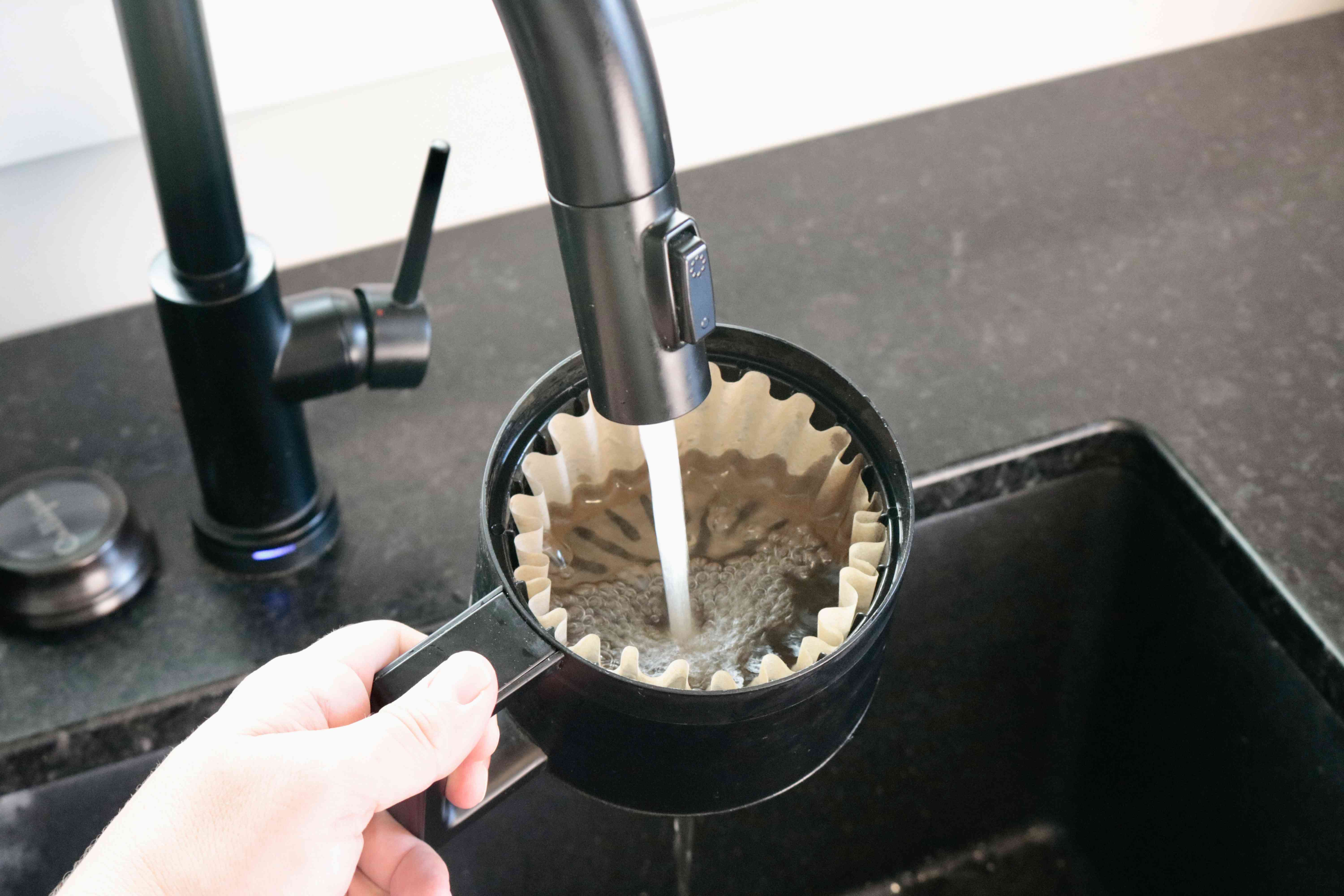 A hand rinsing a coffee placed in a brew basket under the faucet