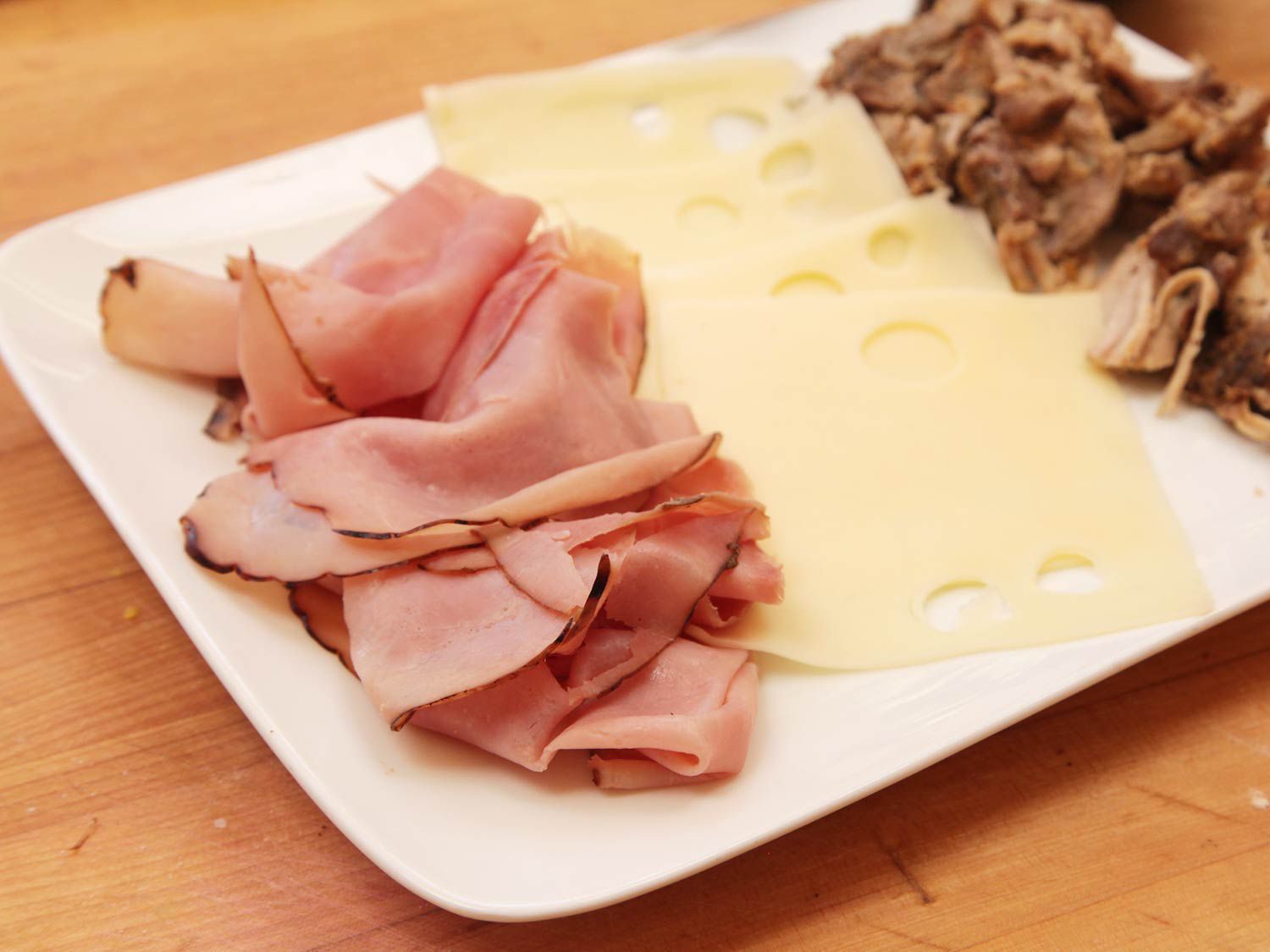 Cuban sandwich ingredients on a white platter: honey ham slices, Swiss cheese, and pulled pork.