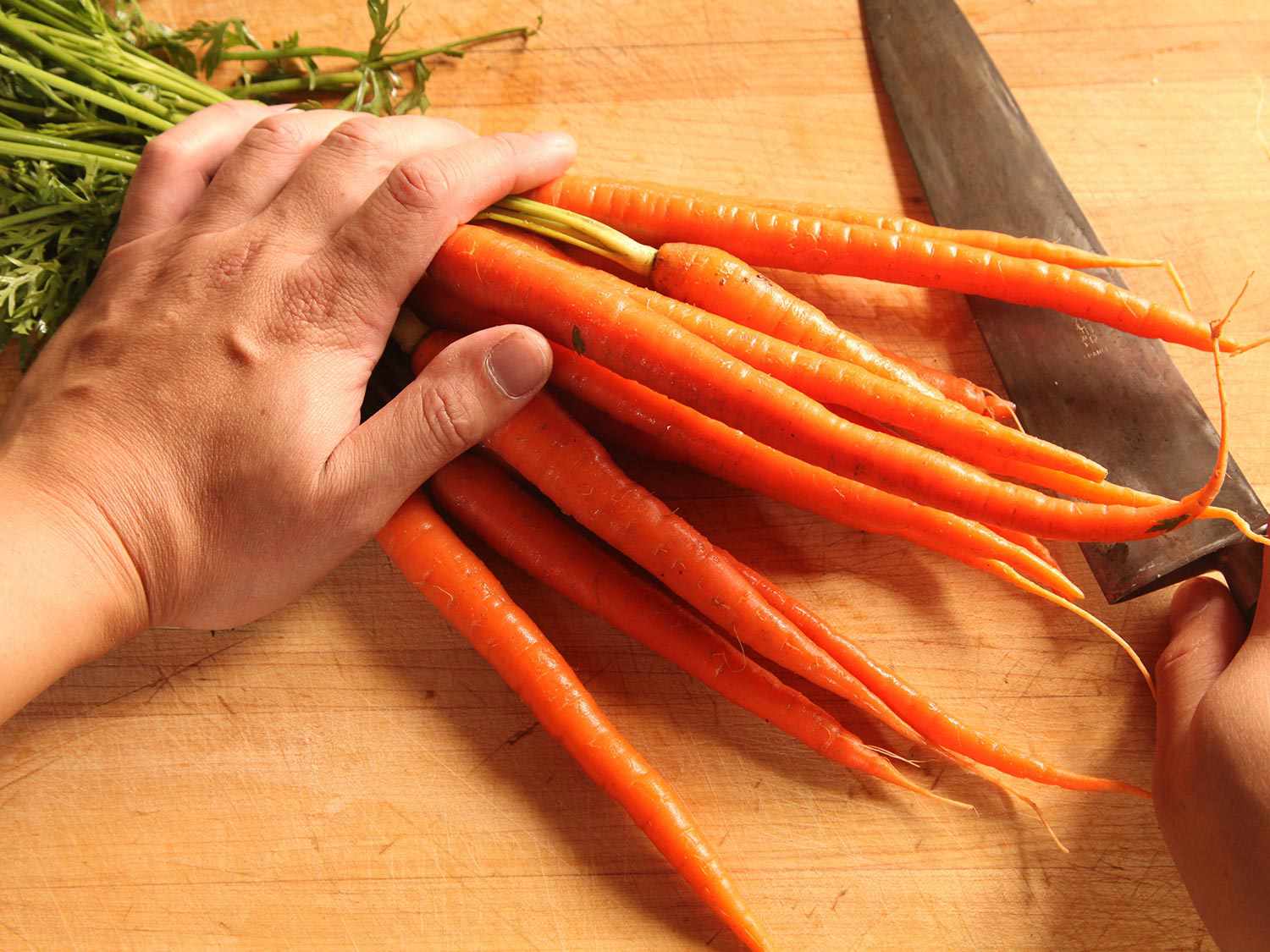 一手把胡萝卜束压在砧板上，另一手拿刀。
