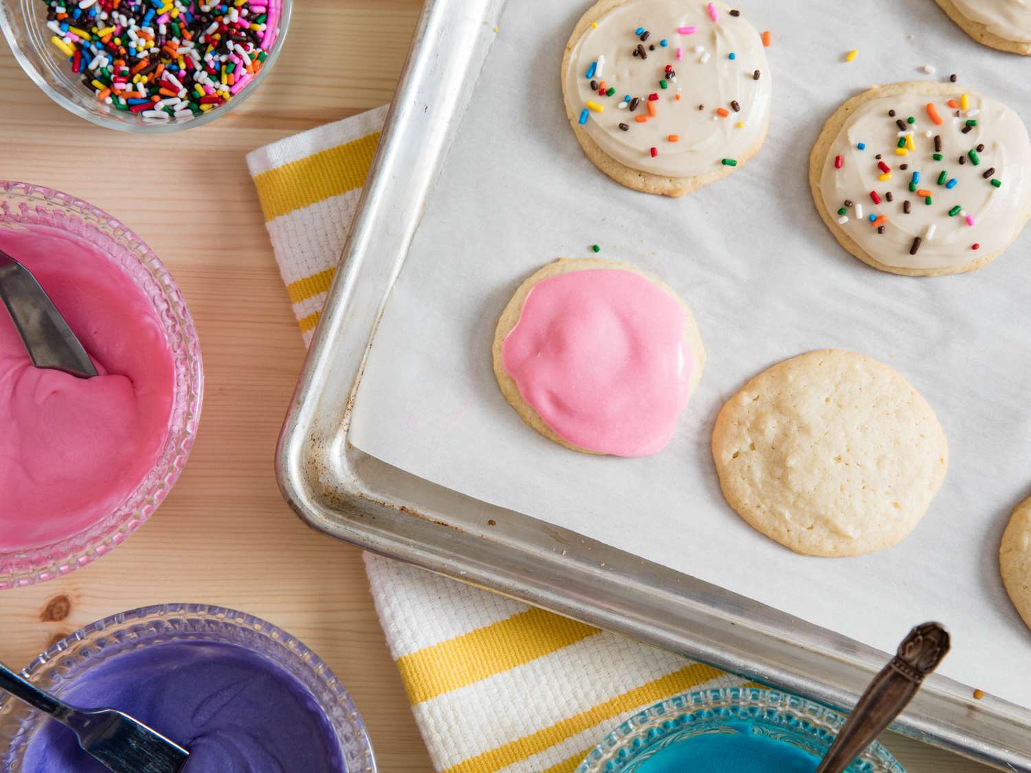Overhead shot of frosted sugar cookies on baking sheet next to bowls of colorful icing.