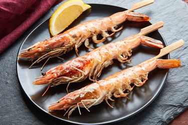 Three cooked head-on shrimp on skewers on a black plate with a lemon wedge.