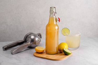 一瓶酸混合木砧板智慧h a lemon, sliced lime, citrus squeezer, and a mixed drink in a glass.