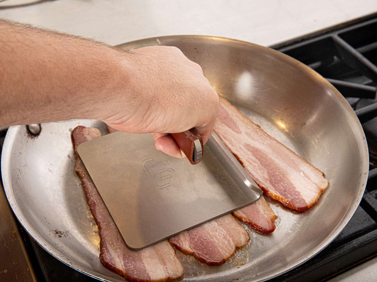 hand using grill press to flatten bacon in a skillet