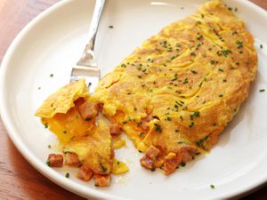 A diner-style ham and cheese omelette on a white plate, sprinkled with chives. A fork is lifting a bit of the omelette.-american-omelet-ham-and-cheese-23.JPG