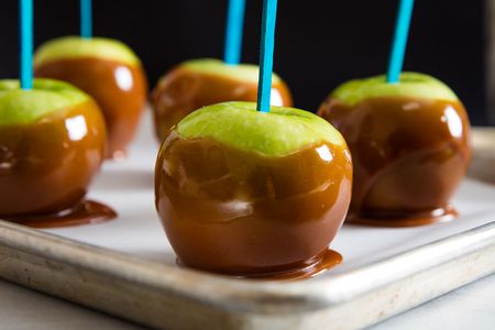 Caramel-dipped Granny Smith apples with blue sticks on a baking tray.