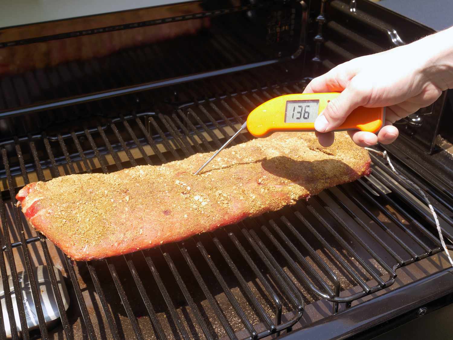 a hand using a thermapen to take the temperature of a rack of ribs on the grill grates.