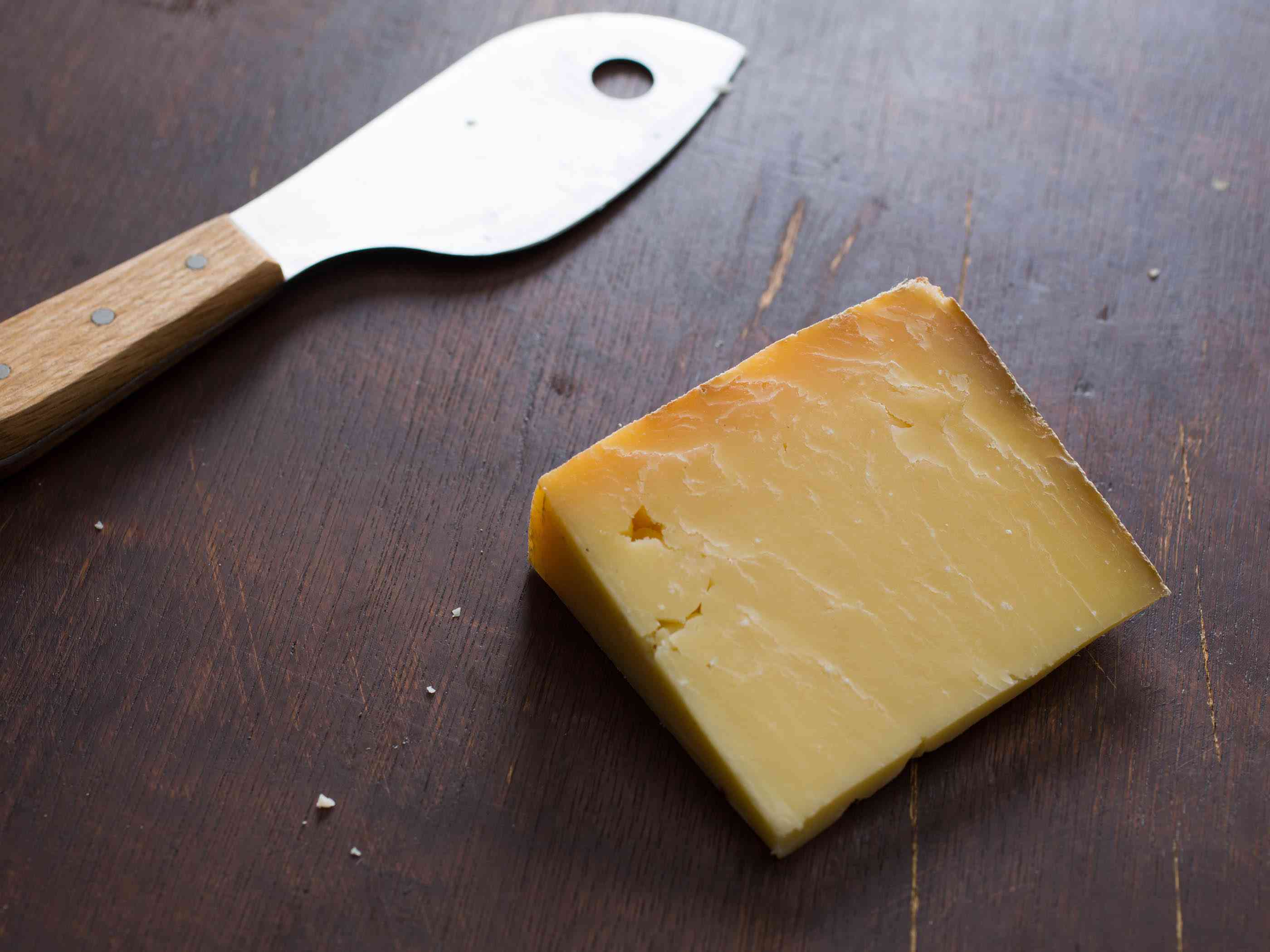 一块楔形的硬奶酪，旁边放一把奶酪刀。它们都放在一张深色的木桌上。