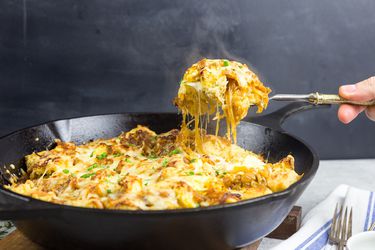French onion strata is served from a cast iron skillet. A portion is lifted from the skillet with a spoon, trailing threads of melted cheese.