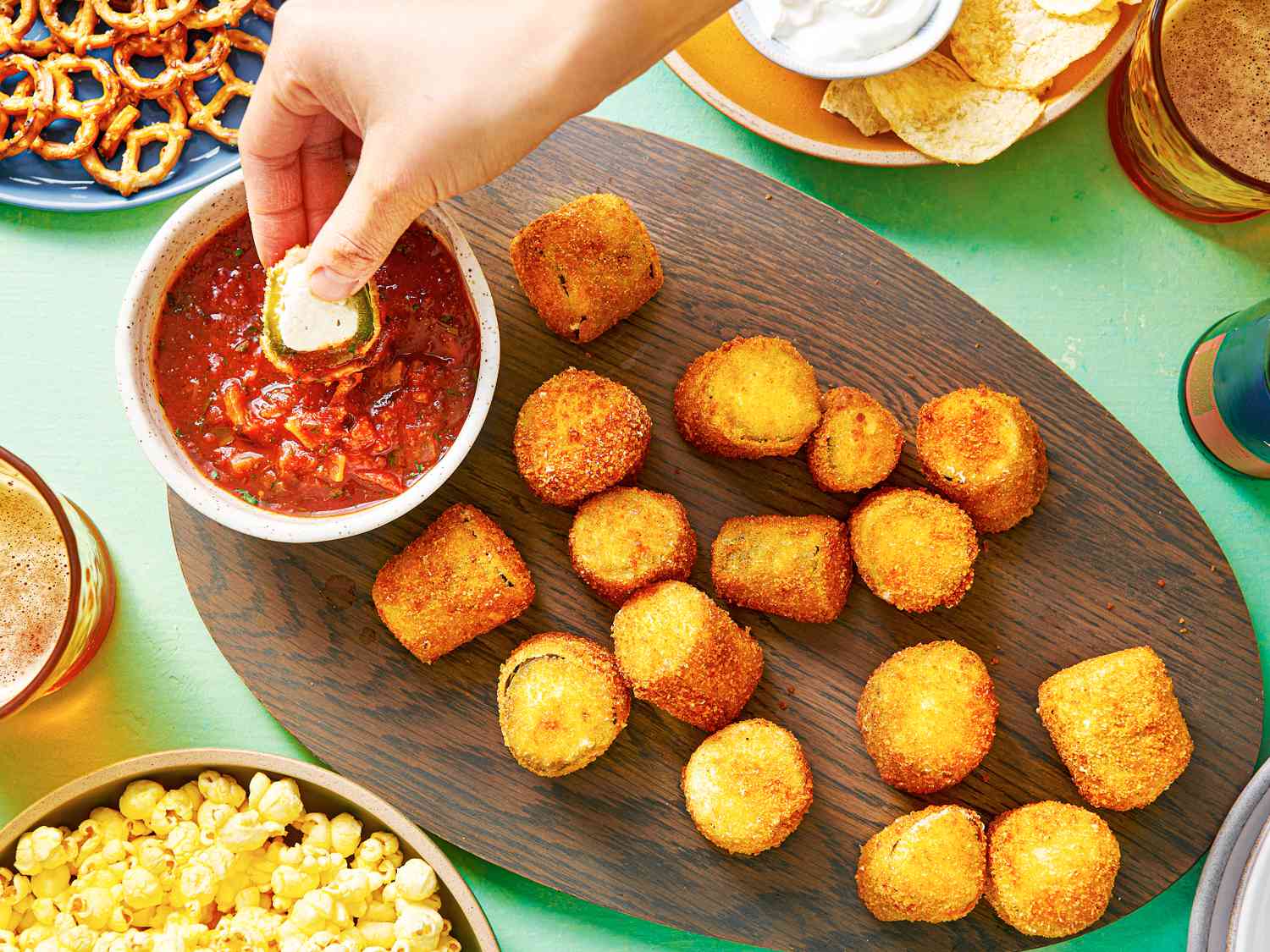 An oval wooden platter holding crispy deep-fried jalapeÃ±o poppers and a small bowl of tomato sauce. There is a hand dipping a popper into the tomato sauce, and the periphery of the image contains other snacks, such as pretzels, popcorn, and chips.