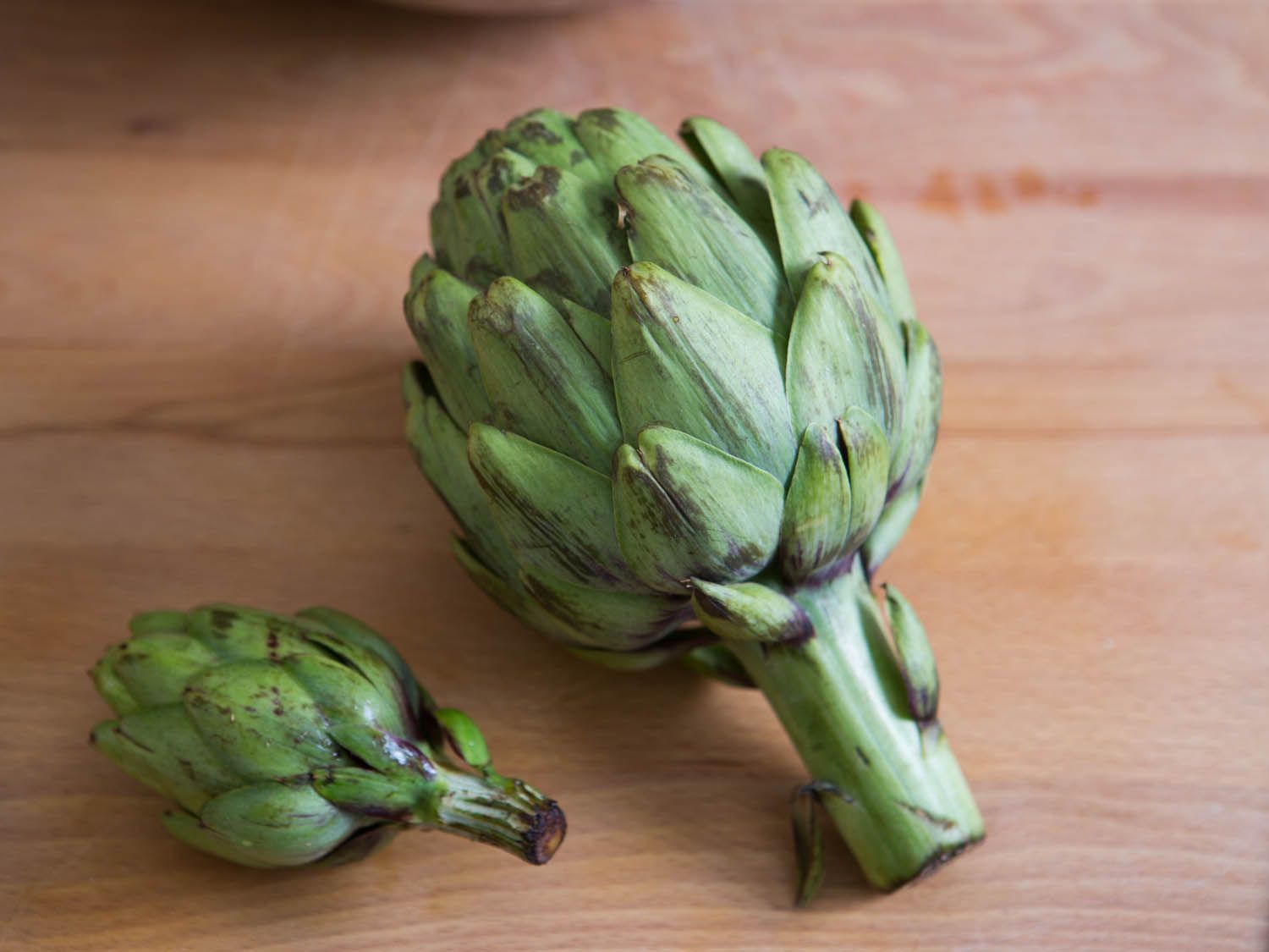 A whole, untrimmed large artichoke next to a whole, untrimmed baby artichoke on a wooden surface.