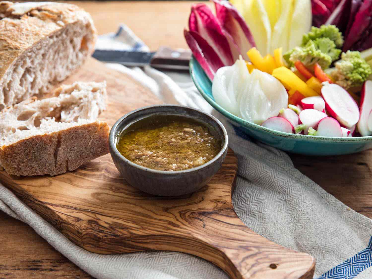 A bowl of bagna càuda dip on a wooden cutting board, with crusty bread and a bowl of colorful assorted vegetables nearby