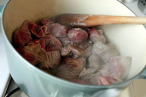 Chunks of beef chuck browning and being stirred in Dutch oven with wooden spoon