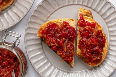 两片面包，上面撒上番茄和洋葱酱，放在奶油色的脊状陶瓷盘子上。图像的左上角是一个装满果酱的玻璃罐，左上角是另一个盘子的一部分，上面有更多的烤面包。