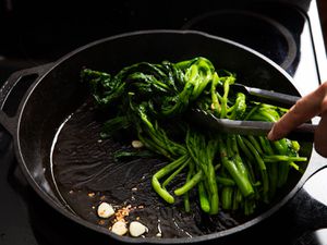 Broccoli rabe being sautéed in a cast iron skillet with a liberal amount of olive oil. Smashed garlic cloves and chile flakes are visible in the pan.