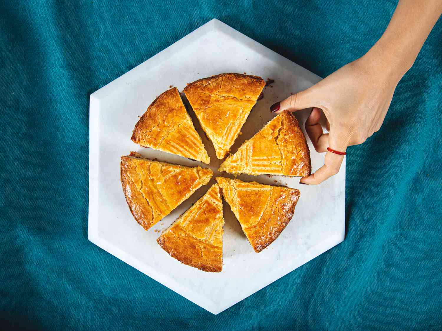 Overhead view of a sliced gateau basque cake with a hand reaching in for a slice