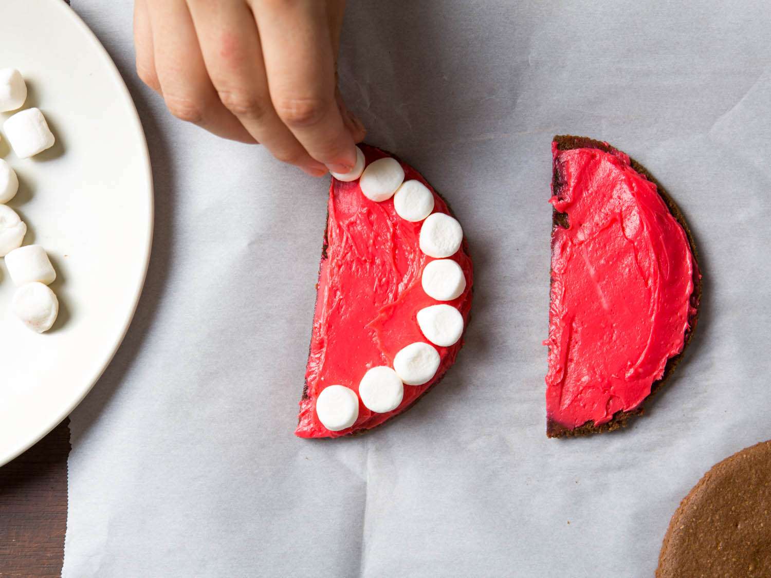 Arranging mini marshmallows along curved edge of chocolate graham cracker half disk iced with red frosting.