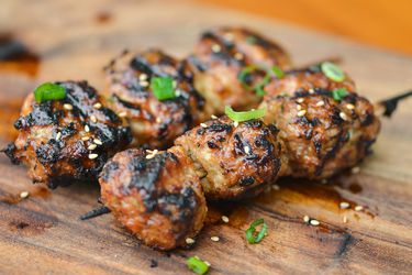 Two skewers of Tsukune, Japanese chicken meatballs, resting on a cutting board and sprinkled with sliced green onions and sesame seeds