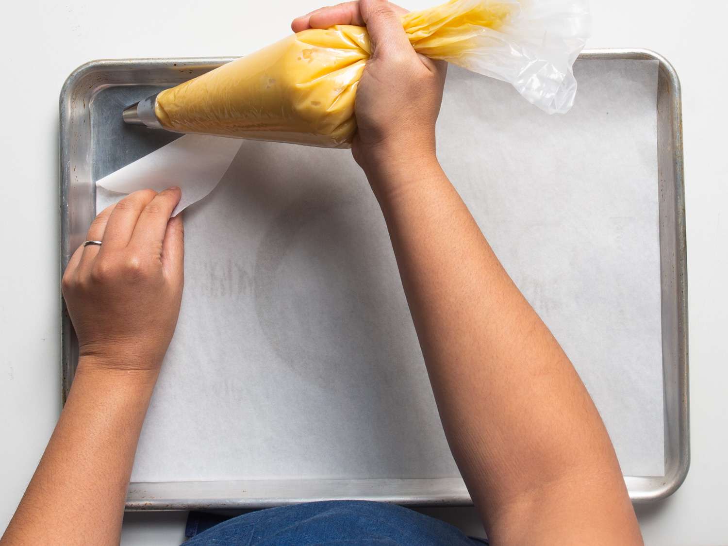 Overhead view of a cook piping a daub of choux paste under a corner of the parchment paper to secure it to the rimmed baking sheet.