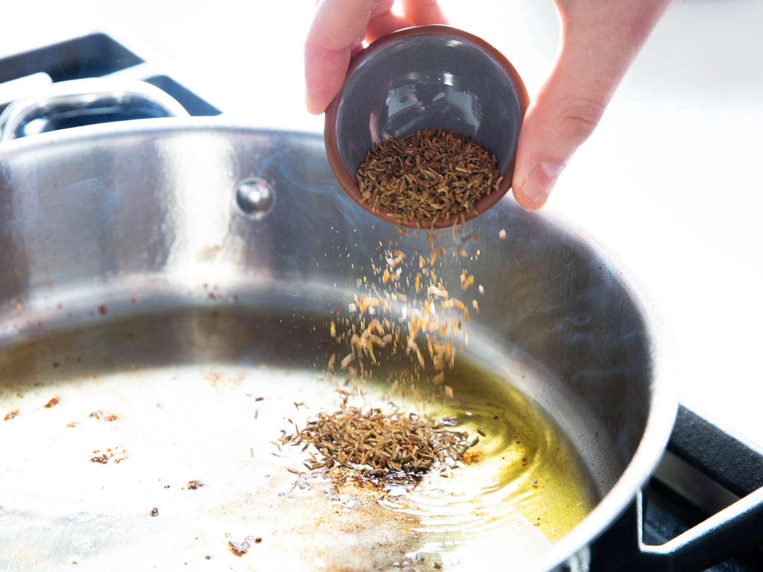 一只手从一个小捏碗里往sauté平底锅里加入孜然种子