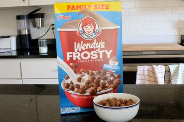 一个box of Wendy's Frosty Cereal (and a bowl of the cereal) atop a black counter.