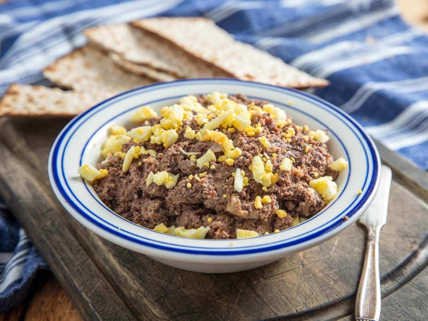 一个博wl of chopped liver topped with minced hard boiled egg and gribenes on a wooden board with matzo.