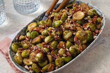 Brussels sprouts in a ceramic serving dish with tongs, a napkin, and glass cups on the periphery