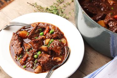 All-American beef stew in a white bowl on a wooden table with a grey Dutch oven of stew in the background.