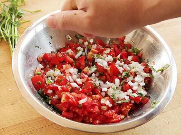 一只手在盛有切碎的西红柿、白洋葱、胡椒和香菜的金属碗上挤酸橙，背景中可以看到多余的香菜茎