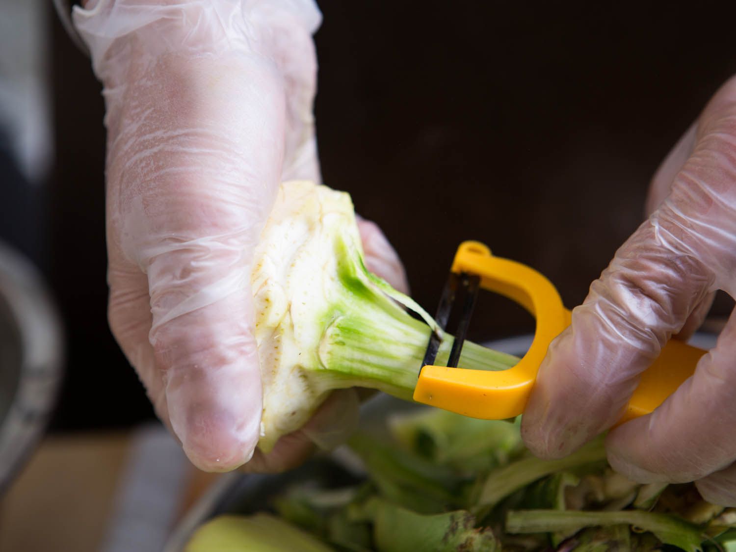 A gloved hand peeling the stem of an artichoke with a yellow Y-peeler.