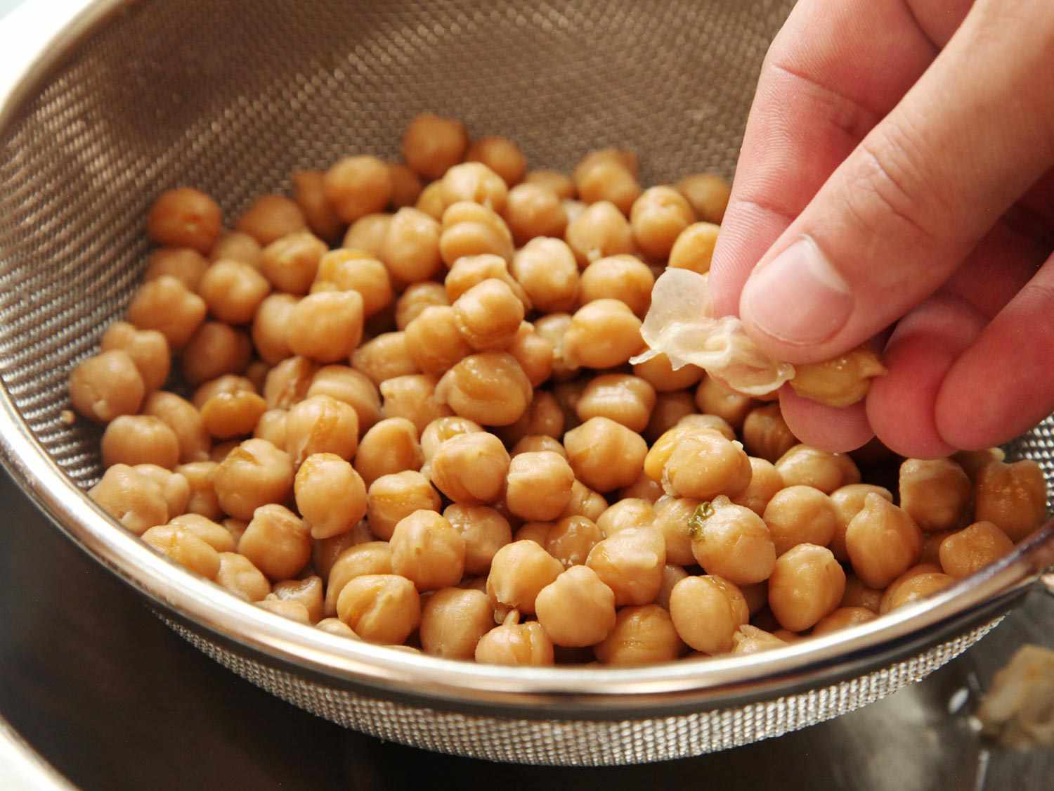 A hand removing papery skin from a chickpea, with a strainer full of chickpeas below