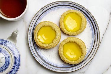 Three Hong Kong egg custard tarts on a plate net to a cup of tea and a tea pot.