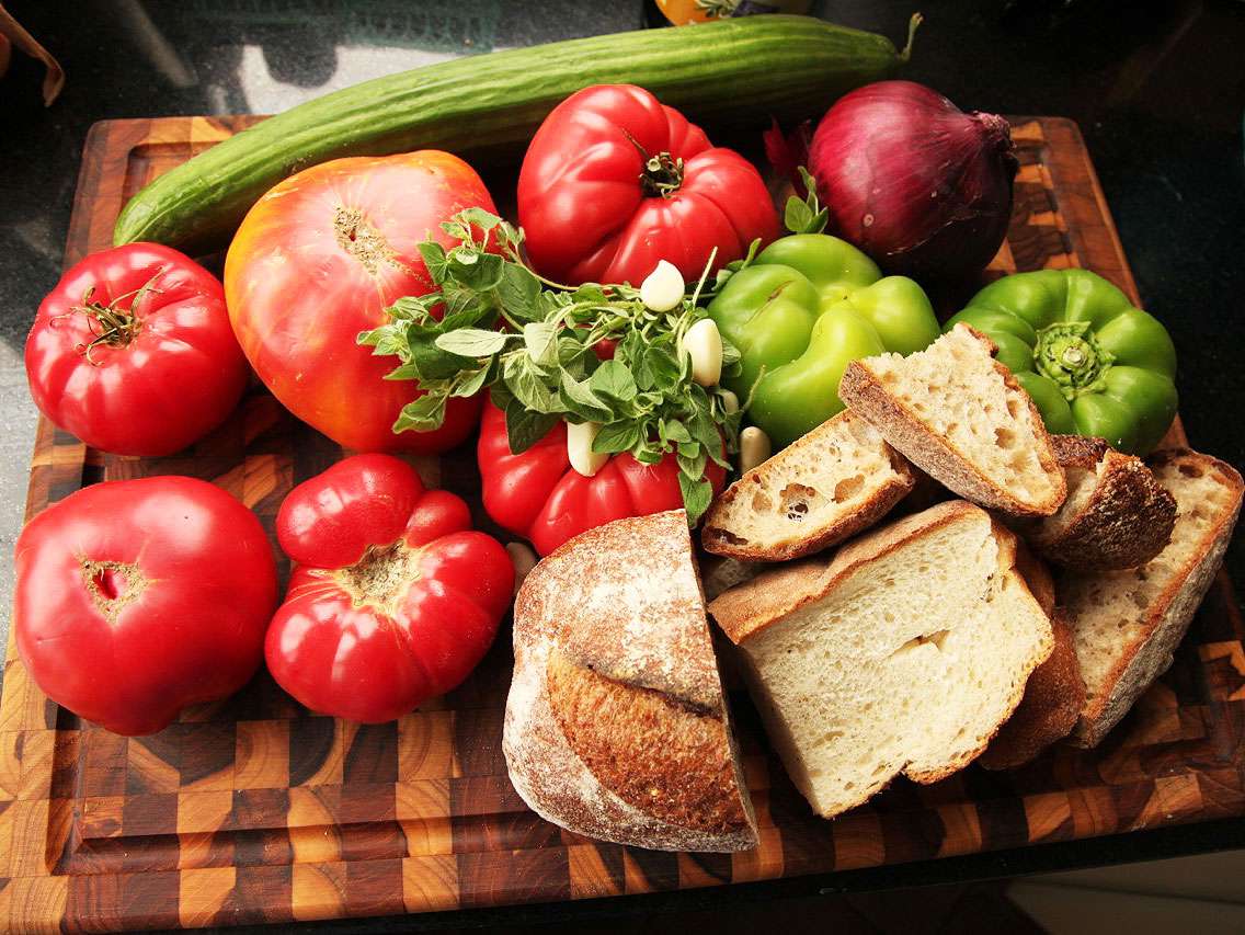 Ingredients for gazpacho on a large wooden cutting board: tomatoes, bread, garlic, oregano leaves, red onions, green bell pepper, and cucumber