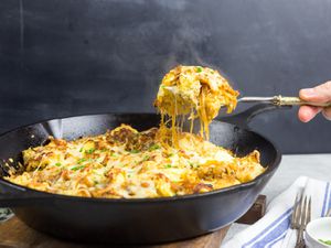 French onion strata is served from a cast iron skillet. A portion is lifted from the skillet with a spoon, trailing threads of melted cheese.
