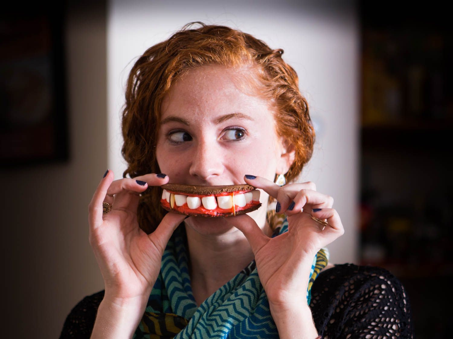Woman holding vampire teeth sandwich cookie in front of her mouth.