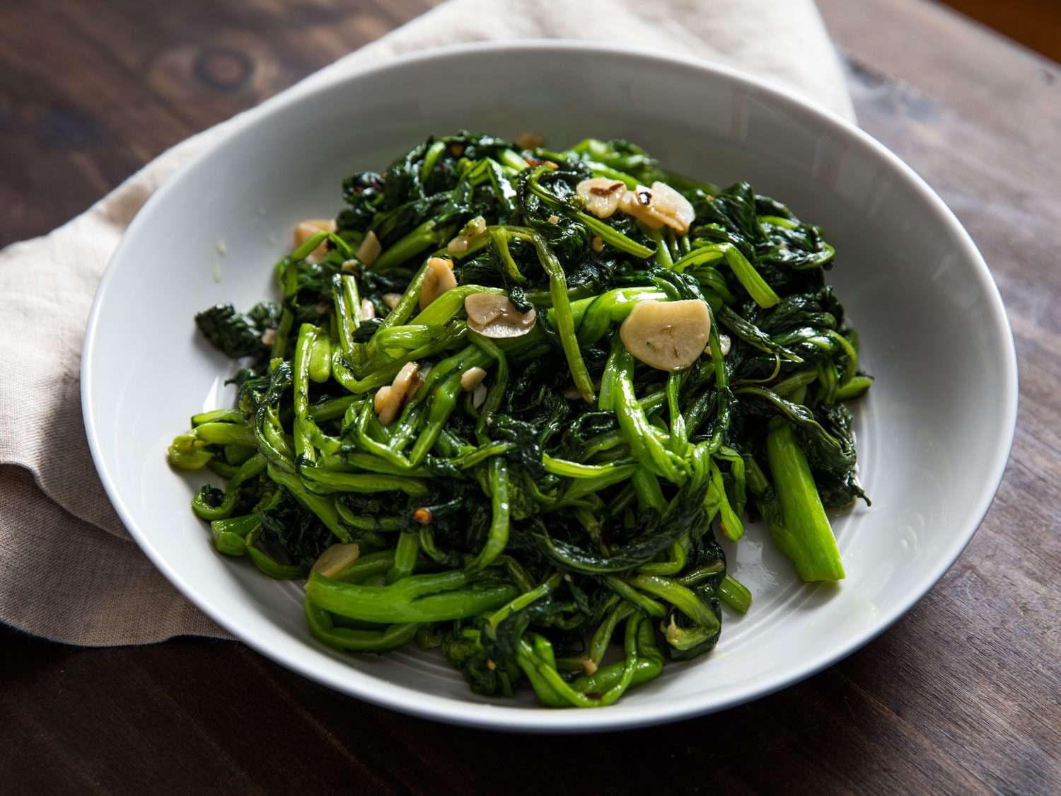 A white bowl of sautéed broccoli rabe, studded with garlic slices and chile flakes.