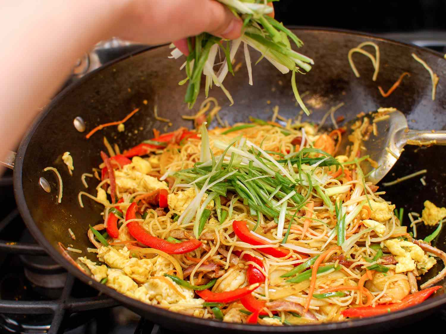 A hand sprinkling sliced scallions into wok of Singapore rice noodles.