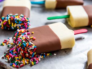 A group of five homemade pudding pops on a baking sheet, dipped in chocolate and covered in candy sprinkles.