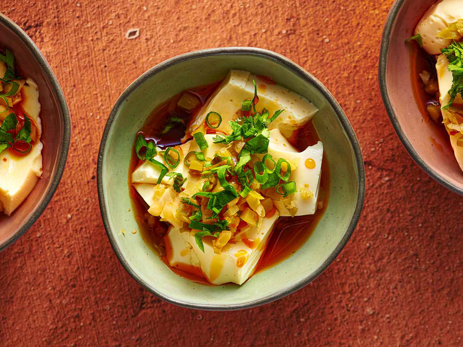 An irregularly round glazed ceramic bowl holding silken tofu with soy sauce and chili oil. There are two additional bowls, one on the right periphery of the image, one on the left periphery.