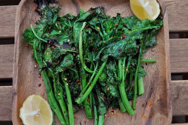 Grilled broccoli rabe with crushed red pepper and lemon wedges on a wooden square plate.