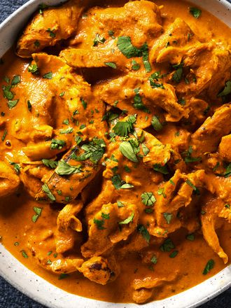 Chicken tikka masala in a white speckled ceramic bowl. In the top left corner is a small bowl holding cilantro leaves and on the top right edge of the image is a plate holding naan bread.