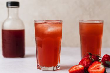 Two glasses of berry shrub. On the right hand side of the image are some strawberries, both whole and sliced in half, and on the left side of the image, in the background, is a small glass bottle holding more shrub.