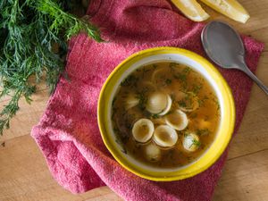 A bowl of chicken noodle soup in a yellow bowl alongside a spoon, fresh dill, and lemon wedges.