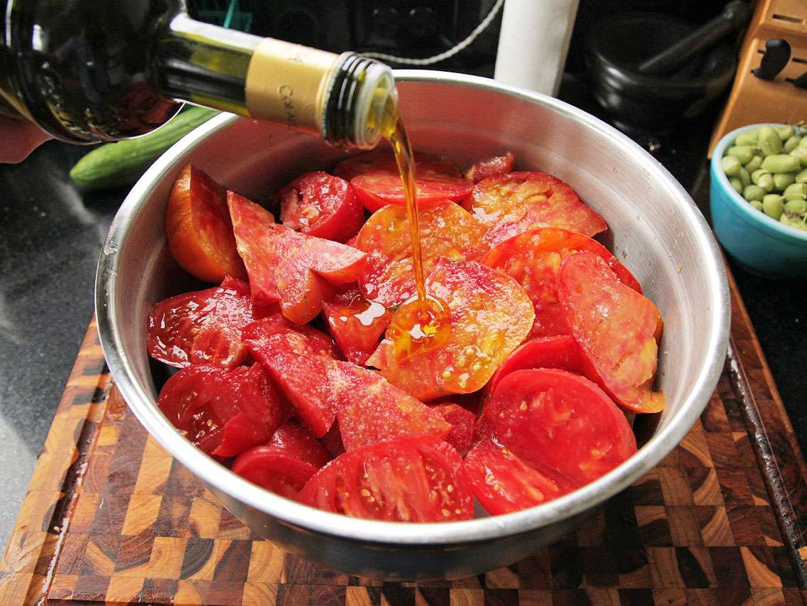 Pouring olive oil over the tomatoes lying on top of torn bread pieces in a large steel mixing bowl for gazpacho.