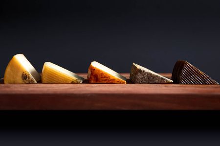 Profile view of five different Manchego-type cheeses, cut into wedges and arranged on a wooden board.