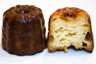 A whole canelé next to another canelé cut open to reveal its custardy interior.