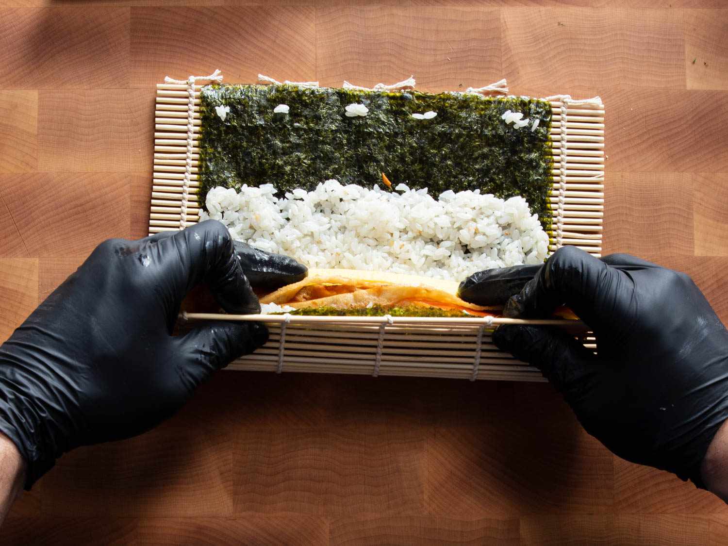 Hands in black gloves rolling up a seaweed roll filled with rice and sliced fillings.