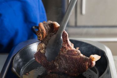 Pair of tongues picking up a piece of meat from cast iron skillet