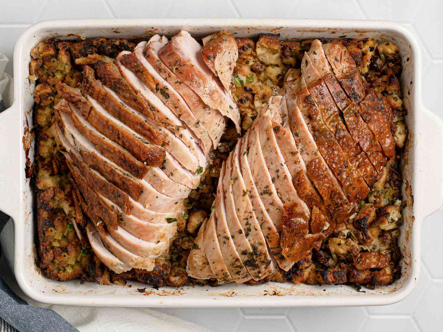 Sliced, herb-roasted turkey breast on top of stuffing inside of a white stoneware baking dish.